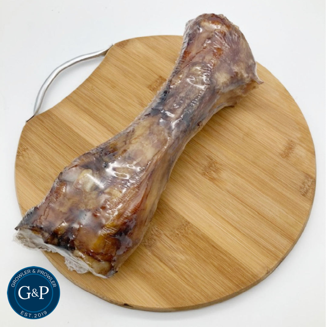 A large, raw beef shin bone resting on a wooden cutting board. The bone is wrapped in plastic and has a light brown colour.